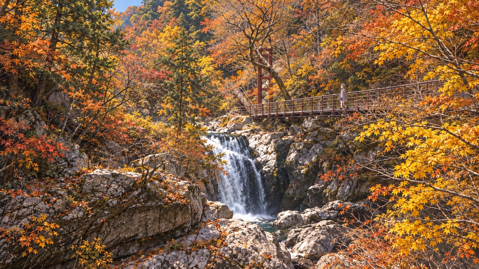 At Mitarai Gorge in autumn, a small figure in a coat stands on the suspension bridge above a waterfall, surrounded by autumn leaves and huge boulders.